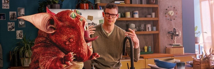 A man standing in his kitchen with an annoyed look as a red creature yells at him